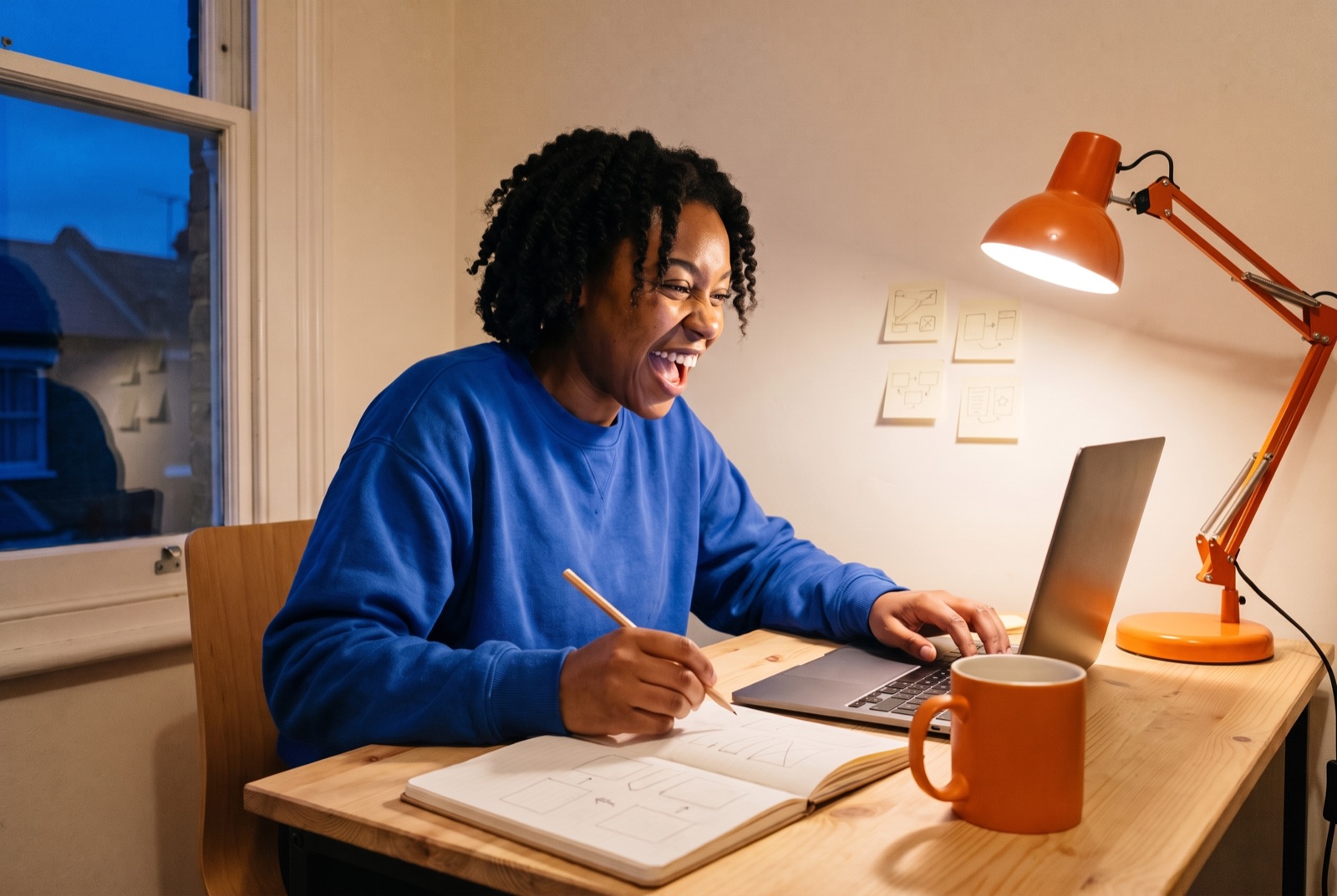 A young woman laughing at her laptop at a desk at night, a notebook of sketches in front of her.