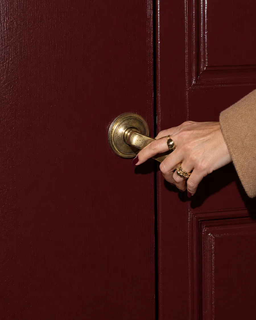 A woman's hand with gold rings pressing down the brass handle of an oxblood front door.